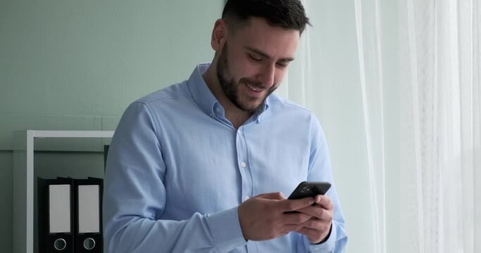 Successful Caucasian businessman is standing next to a window in his office, looking at his phone and typing on it. Suddenly, he starts laughing, showing his white teeth and a wide smile.
