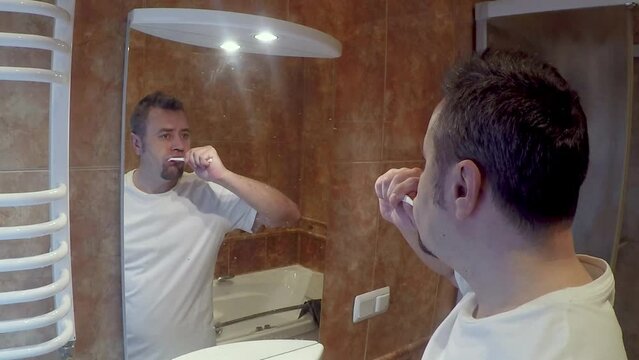 Young man brushes his teeth in the bathroom in front of a mirror