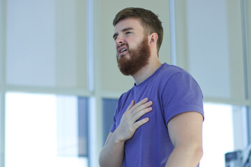 Tired exhausted young man is training workout, running on treadmill in gym holding his heart with...