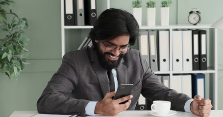 Slow-motion footage, an Indian businessman sits in his office texting while sipping a hot beverage from a mug. Suddenly, he receives a funny message that makes him burst out laughing with joy.