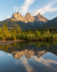 Naklejka na ściany i meble Three Sisters in Canmore seen at golden hour, sunset on blue sky day, afternoon with calm, peaceful reflection in water below famous, tourist, tourism mountains, area summer. 