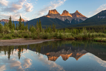 Three Sisters in Canmore seen at golden hour, sunset on blue sky day, afternoon with calm, peaceful reflection in water below famous, tourist, tourism mountains, area summer.  