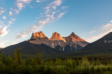 Naklejka premium Sunset wilderness views in Canmore during summer with Three Sisters in view. Golden glow on mountain peaks. Background, wallpaper, desktop scenic view, tourist, tourism area. Blue sky, clouds