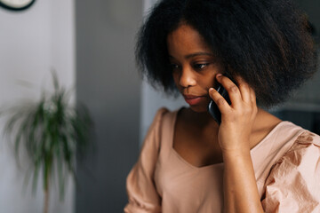 Closeup face of thoughtful young African American woman talking on smartphone at home. Close up portrait of attractive black female calling mobile phone in apartment. People and technology concept.