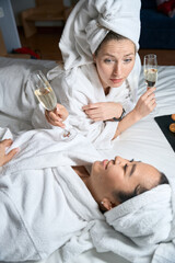 Two women in white bathrobes talking while lying on bed