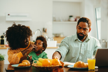 Fototapeta na wymiar An african american mother is feeding her son while sitting with her working father at the breakfast table at home in the morning.