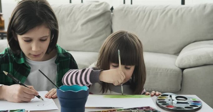 A Sister and brother seated near a table on the floor, they dive into their creative pursuits, skillfully using pencils, paintbrushes, and watercolors.