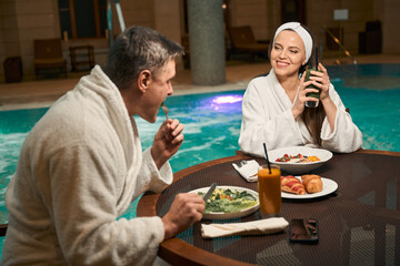 Fototapeta na wymiar Mature couple in bathrobes having lunch by swimming pool