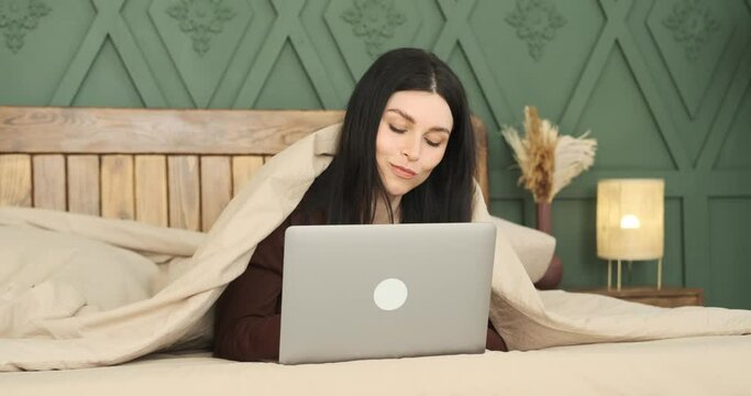 A serene Caucasian woman in cozy pajamas lies peacefully on her bed, embraced by a warm blanket. With a genuine smile and thoughtful eyes, she engrosses herself in a captivating laptop conversation.