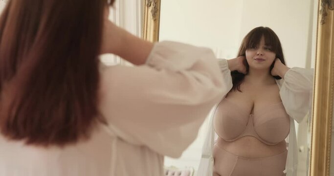 Plus-size Caucasian woman in a robe and lingerie. In the comfort of her bedroom, she stands gracefully by the mirror, gazing at her reflection with an air of contentment and positivity.