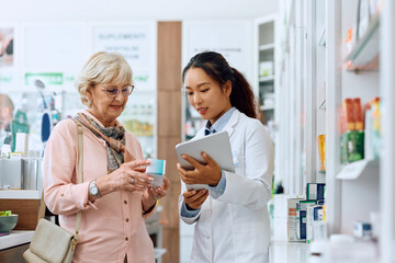 Chinese pharmacist and her senior customer using touchpad while searching for medicine in drugstore.