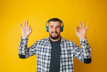 Naklejka premium Happy caucasian guy shows a good gesture, agrees with something, claims that everything is fine, smiles broadly, wears headphones, enjoys isolated on a yellow background