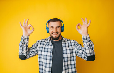 Naklejka premium Happy caucasian guy shows a good gesture, agrees with something, claims that everything is fine, smiles broadly, wears headphones, enjoys isolated on a yellow background