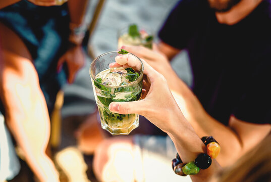 Refreshing Summer Mojito - Close-up of a hand holding a vibrant mojito cocktail with flames on ice. Friends enjoy the lively atmosphere in the background. Ideal for summer-themed projects.