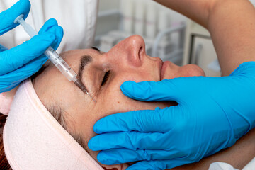 The beautician injects hyaluronic acid into the client's cheek, filling the wrinkles around the...
