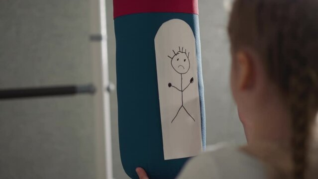 Schoolgirl draws upset man on punching bag in playing room close rare view. Little girl visualizes anger feeling to deal with offence in home gym