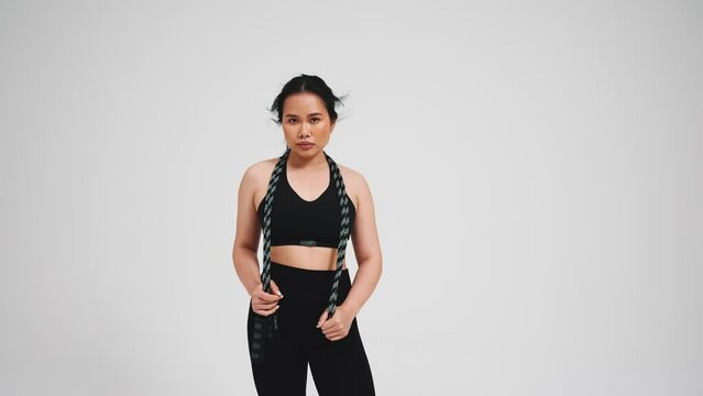 Pretty asian woman holding skipping rope in black tracksuit against gray background in studio 