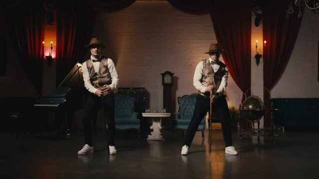 Two stylish professional male dancers in classy dandy hat performing jazz funk dance synchronic moves in vintage production studio retro environment with theatrical red velvet curtains and candlelight