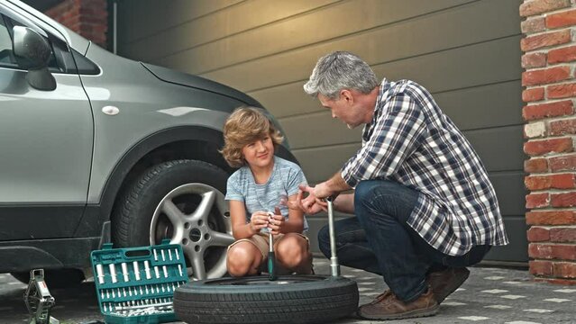 Cute little boy helps father to change wheel on their family car on warm day in the yard. Male family members siting next to car and communicate with each other. Son and dad spending time together.