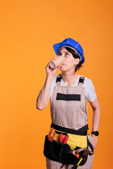 Construction worker serving coffee cup after renovation, using building tools to restore and decorate. Professional contractor wearing protective helmet and overalls in studio against yellow