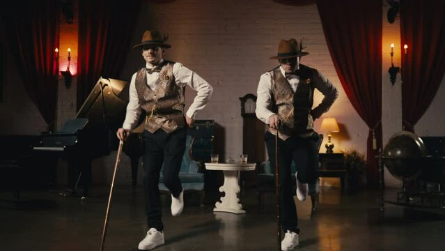Two guys in dandy style outfit synchronically dancing in retro theatre stage. Classy men dancers performing in jazz funk style, dancing with classic cane in hands. Music video, night show or event 4K