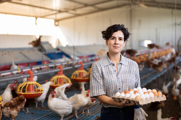 Happy successful hispanic female owner of poultry farm engaged in breeding of laying hens standing in henhouse, holding cardboard tray of fresh organic chicken eggs - obrazy, fototapety, plakaty