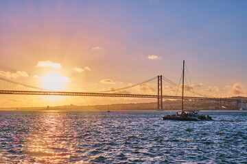 Fototapeta premium View of 25 de Abril Bridge famous tourist landmark of Lisbon connecting Lisboa and Almada on Setubal Peninsula over Tagus river with tourist yacht silhouette at sunset. Lisbon, Portugal