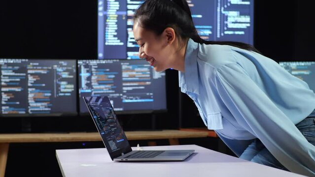 Side View Of Asian Female Programmer Walking Into The Working Space To Write Code By A Laptop Using Multiple Monitors Showing Database On Terminal Window Desktops In The Office
