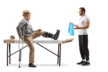 Elderly male patient sitting on a therapy bed with a brace on his leg and a therapist showing...