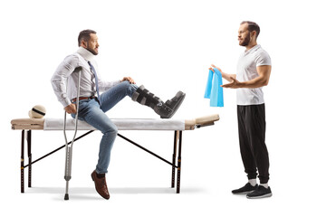 Obraz premium Young male patient sitting on a therapy bed with a brace on his leg and a therapist showing exercises with a resistance band