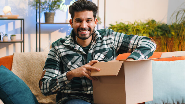 Happy indian man unpacking delivery parcel sitting at home apartment. Smiling satisfied hindu guy shopper, online shop customer opening cardboard box receiving purchase gift by fast postal shipping