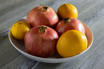 Three of fresh ripe pomegranates and three oranges in a plate on a gray wooden table. Close-up