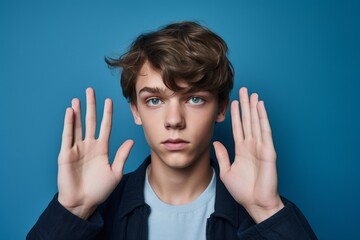 Headshot portrait photography of a tender boy in his 20s making a sorry gesture with hands together against a sapphire blue background. With generative AI technology
