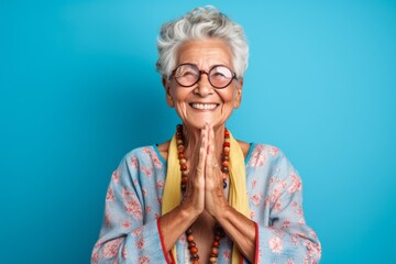 Headshot portrait photography of a happy old woman joining palms in a gesture of gratitude against a cerulean blue background. With generative AI technology
