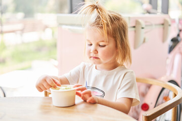 Cute little girl is getting ready to try ice cream. A beautiful charming baby looks to the side sitting at a table in a cafe near the window. Children's cafe. The interior of an ice cream cafe.