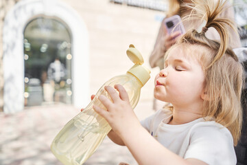a charming little girl drinks water from a sports bottle in a city park. a close-up portrait of a...