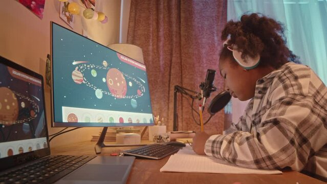 Waist up of African American gen z girl listening to music in wireless headphones and drawing while sitting at desk by computer with planets of solar system on wallpaper at night