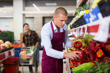 Supermarket worker stacking red peppers on shelf in salesroom. Woman with cart shopping in background.