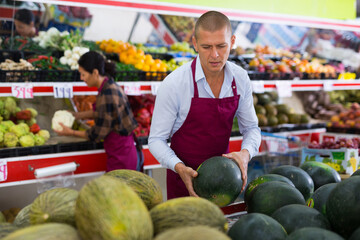 Supermarker worker in apron standing in salesroom among shelves with fruits and vegetables and...