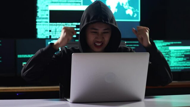 Asian Female Hacker In A Hood Celebrating Succeed Attack Global Infrastructure Servers By A Laptop With Maps And Data On Display Screens Of Multiple Monitors In A Dark Office Room
