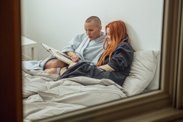 A couple of women in love are reading a book in bed. View through the window. Relationships in an LGBT couple.