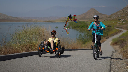 Mother and daughter biking up a hill next to a lake with mountains riding recumbent and normal...