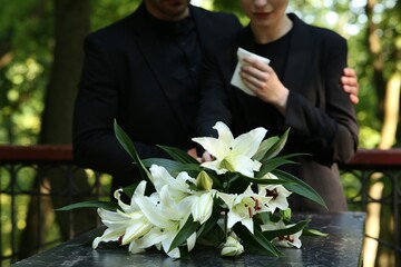 Couple near granite tombstone with white lilies at cemetery outdoors, selective focus. Funeral...