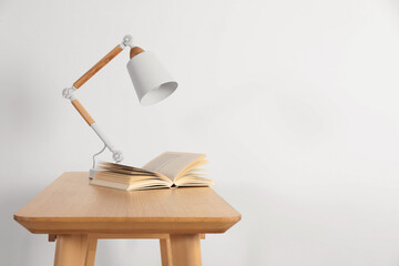 Stylish modern desk lamp and open book on wooden table near white wall, space for text