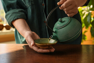 Woman pouring freshly brewed tea from teapot into cup at wooden table indoors, closeup. Traditional...