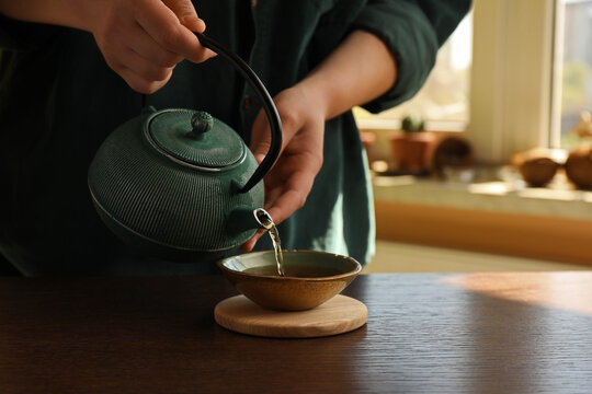 Woman pouring freshly brewed tea from teapot into cup at wooden table indoors, closeup and space for text. Traditional ceremony