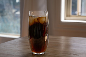 A clear tall glass of dark cola with multiple iceberg ice cubes on a wooden table. The drink has...