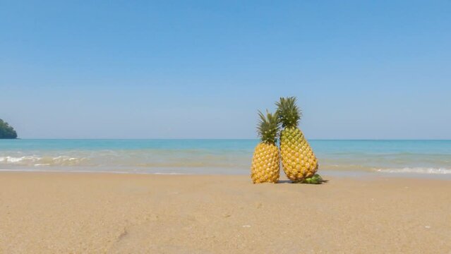 two pineapples on the fine sandy beach in blue clear sky with blue ocean sea. foaming waves crashing on the sand. traveling on holiday vacation on a sunny summer day to paradise. sunshine on sand.