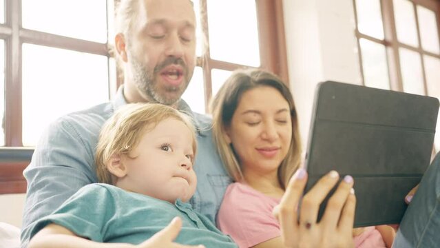 Caucasian parents and little baby son resting on the bed in bedroom with using digital tablet play learning games or watch movie together. Happy family enjoy weekend activity with technology at home.