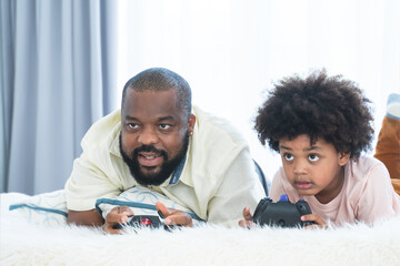 African father and child son playing online video games, using joysticks or game console, having...
